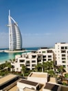 A modern hotel building with a unique sail-like design is set against a clear blue sky. In the foreground, there is a luxurious resort complex with cream-colored buildings surrounded by lush palm trees and well-maintained gardens. The turquoise sea provides a stunning backdrop, enhancing the serene and upscale ambiance of the area.