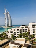 A modern hotel building with a unique sail-like design is set against a clear blue sky. In the foreground, there is a luxurious resort complex with cream-colored buildings surrounded by lush palm trees and well-maintained gardens. The turquoise sea provides a stunning backdrop, enhancing the serene and upscale ambiance of the area.