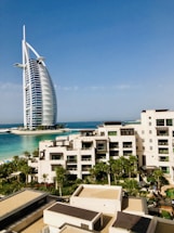 A modern hotel building with a unique sail-like design is set against a clear blue sky. In the foreground, there is a luxurious resort complex with cream-colored buildings surrounded by lush palm trees and well-maintained gardens. The turquoise sea provides a stunning backdrop, enhancing the serene and upscale ambiance of the area.