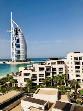 A modern hotel building with a unique sail-like design is set against a clear blue sky. In the foreground, there is a luxurious resort complex with cream-colored buildings surrounded by lush palm trees and well-maintained gardens. The turquoise sea provides a stunning backdrop, enhancing the serene and upscale ambiance of the area.