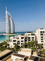 A modern hotel building with a unique sail-like design is set against a clear blue sky. In the foreground, there is a luxurious resort complex with cream-colored buildings surrounded by lush palm trees and well-maintained gardens. The turquoise sea provides a stunning backdrop, enhancing the serene and upscale ambiance of the area.