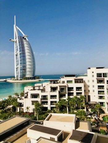 A modern hotel building with a unique sail-like design is set against a clear blue sky. In the foreground, there is a luxurious resort complex with cream-colored buildings surrounded by lush palm trees and well-maintained gardens. The turquoise sea provides a stunning backdrop, enhancing the serene and upscale ambiance of the area.