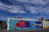 A vibrant mural featuring a large, colorful fish with scales transitioning from pink and purple to blue, set against a bright turquoise background. The fish incorporates images of marine life and ocean elements. The scene is outdoors, with a clear blue sky and a boardwalk in the foreground. In the background, some urban buildings are visible.