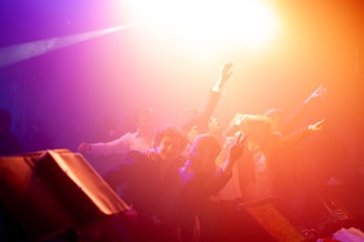 A group of people is celebrating energetically under vibrant lighting. The scene is filled with bright colors, suggesting a lively party atmosphere. Individuals appear to be dancing, with some holding drinks, and the light creates a dramatic effect.