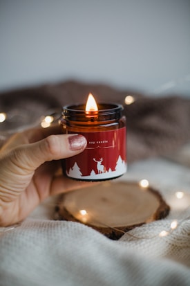 A hand with painted fingernails holds a lit soy candle in a red jar with white silhouettes of trees and a deer. The background is soft, featuring a wooden disc and glowing string lights.