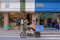 A street scene with people interacting at a small storefront that combines a cafe and a clothing store. The cafe features a service window with patrons, while the clothing store displays garments visible through a glass facade. A cyclist on a motorized bike with a large blue basket is passing by in the foreground.