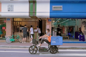 A street scene with people interacting at a small storefront that combines a cafe and a clothing store. The cafe features a service window with patrons, while the clothing store displays garments visible through a glass facade. A cyclist on a motorized bike with a large blue basket is passing by in the foreground.