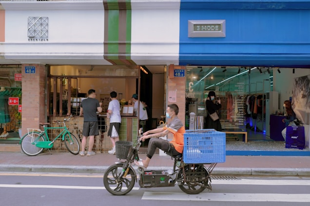 A street scene with people interacting at a small storefront that combines a cafe and a clothing store. The cafe features a service window with patrons, while the clothing store displays garments visible through a glass facade. A cyclist on a motorized bike with a large blue basket is passing by in the foreground.