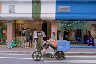 A street scene with people interacting at a small storefront that combines a cafe and a clothing store. The cafe features a service window with patrons, while the clothing store displays garments visible through a glass facade. A cyclist on a motorized bike with a large blue basket is passing by in the foreground.