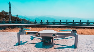 A compact drone is resting on a flat, stone surface. In the background, a scenic view of distant mountains is visible under a clear blue sky. A railing and some greenery line the horizon, suggesting an elevated location.