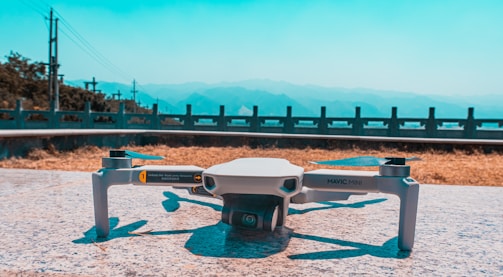 A compact drone is resting on a flat, stone surface. In the background, a scenic view of distant mountains is visible under a clear blue sky. A railing and some greenery line the horizon, suggesting an elevated location.