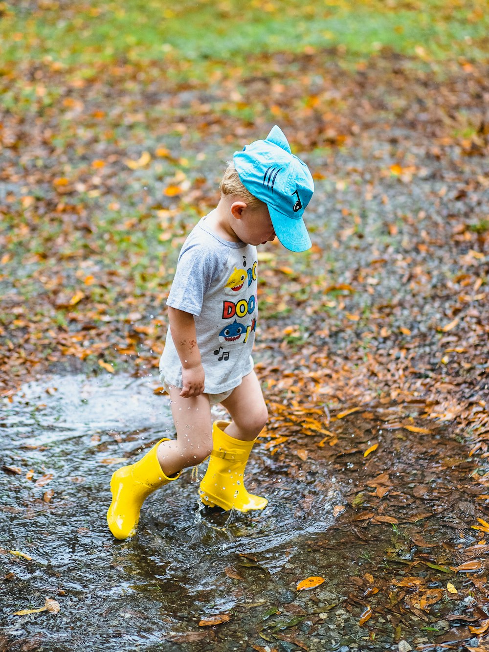child in white t-shirt and yellow rain boots walking on brown dried leaves during daytime