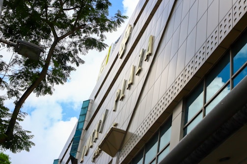 View of a tall building facade with large windows reflecting the sky. A tree branch and streetlight appear in the foreground, casting shadows on the structure. The architectural design features perforated metal panels and a modern aesthetic.