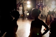 Two silhouetted figures of dancers are in the foreground, likely backstage, facing each other amidst a dimly lit environment. Bright stage lights illuminate the background, revealing a group of performers in costumes, suggesting a theatrical or ballet performance setting.
