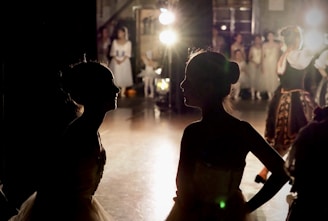 Two silhouetted figures of dancers are in the foreground, likely backstage, facing each other amidst a dimly lit environment. Bright stage lights illuminate the background, revealing a group of performers in costumes, suggesting a theatrical or ballet performance setting.