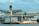 A small boat filled with people is docked at a floating fuel station named Petrobras. The station is on a body of water, adjacent to some industrial and warehouse buildings. The boat is painted white and yellow, with a canopy protecting some passengers. The background features a cloudy sky and structures with metallic roofs.