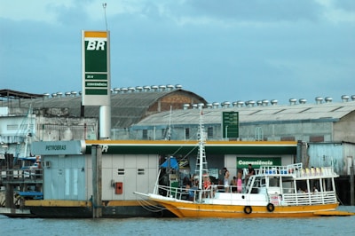 A small boat filled with people is docked at a floating fuel station named Petrobras. The station is on a body of water, adjacent to some industrial and warehouse buildings. The boat is painted white and yellow, with a canopy protecting some passengers. The background features a cloudy sky and structures with metallic roofs.