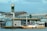A small boat filled with people is docked at a floating fuel station named Petrobras. The station is on a body of water, adjacent to some industrial and warehouse buildings. The boat is painted white and yellow, with a canopy protecting some passengers. The background features a cloudy sky and structures with metallic roofs.