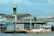 A small boat filled with people is docked at a floating fuel station named Petrobras. The station is on a body of water, adjacent to some industrial and warehouse buildings. The boat is painted white and yellow, with a canopy protecting some passengers. The background features a cloudy sky and structures with metallic roofs.
