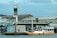 A small boat filled with people is docked at a floating fuel station named Petrobras. The station is on a body of water, adjacent to some industrial and warehouse buildings. The boat is painted white and yellow, with a canopy protecting some passengers. The background features a cloudy sky and structures with metallic roofs.