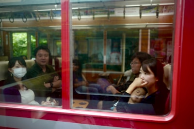 Passengers are seated inside a train or metro carriage. Several people are visible through the window. One woman is wearing a mask, and another woman appears to be deep in thought. A child is sleeping next to her.