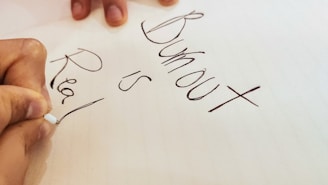 A close-up view of a hand holding a pen, writing the words 'Burnout is Real' on a piece of paper. The handwriting is in black ink, and the background is a cream-colored sheet.