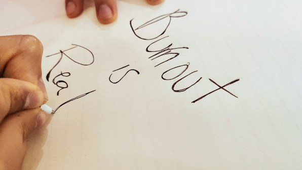 A close-up view of a hand holding a pen, writing the words 'Burnout is Real' on a piece of paper. The handwriting is in black ink, and the background is a cream-colored sheet.