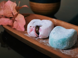 Two mochi desserts rest on a wooden tray, one pink with a chocolate filling partially visible and the other blue, both covered in a light dusting of powdered sugar. A branch of pink bougainvillea flowers is placed beside the mochi. In the background, a dark cup with a wooden texture is partially visible.