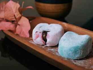 Two mochi desserts rest on a wooden tray, one pink with a chocolate filling partially visible and the other blue, both covered in a light dusting of powdered sugar. A branch of pink bougainvillea flowers is placed beside the mochi. In the background, a dark cup with a wooden texture is partially visible.