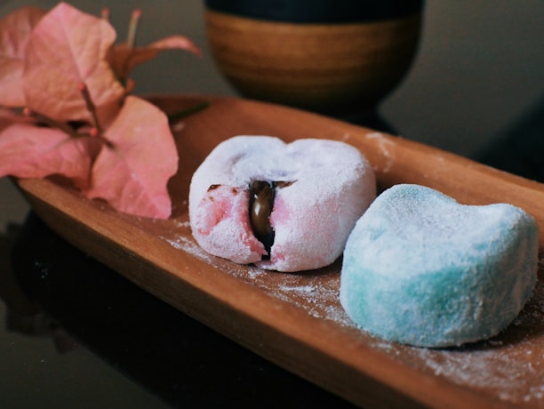 Two mochi desserts rest on a wooden tray, one pink with a chocolate filling partially visible and the other blue, both covered in a light dusting of powdered sugar. A branch of pink bougainvillea flowers is placed beside the mochi. In the background, a dark cup with a wooden texture is partially visible.