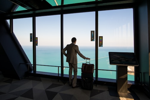 A person in a suit stands in a modern building, looking out through large windows at a vast body of water and sky. The individual has a wheeled suitcase and is positioned near a digital kiosk.