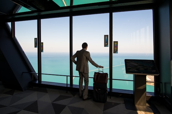 A person in a suit stands in a modern building, looking out through large windows at a vast body of water and sky. The individual has a wheeled suitcase and is positioned near a digital kiosk.