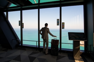 A person in a suit stands in a modern building, looking out through large windows at a vast body of water and sky. The individual has a wheeled suitcase and is positioned near a digital kiosk.