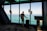 A person in a suit stands in a modern building, looking out through large windows at a vast body of water and sky. The individual has a wheeled suitcase and is positioned near a digital kiosk.
