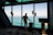 A person in a suit stands in a modern building, looking out through large windows at a vast body of water and sky. The individual has a wheeled suitcase and is positioned near a digital kiosk.