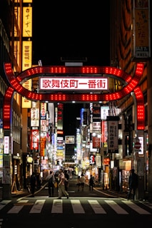 A bustling street at night is illuminated by an array of colorful neon lights and signs written in Japanese. The atmosphere is lively with people walking on the pedestrian crossing and along the sidewalks. Buildings adorned with bright advertisements create a vibrant cityscape.