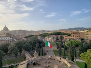 An urban landscape with historic architecture and lush greenery dominates the scene. A prominent Italian flag waves in the foreground, situated atop a fortress-like structure. The background features a cityscape with buildings showcasing classical European design, and a large dome is visible, silhouetted against a clear, blue sky.