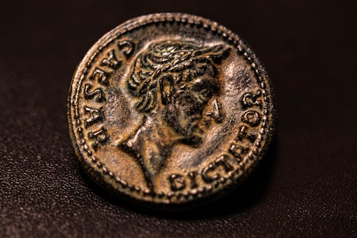 A detailed close-up of an ancient coin with a profile of a figure inscribed with the words 'CAESAR DICTATOR'. The coin displays a worn, textured surface with intricate designs and lettering around the edge.