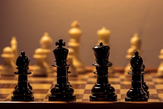 A close-up view of a chessboard highlighting the black chess pieces in the foreground with the white pieces in soft focus in the background. The black pieces, including the king and queen, are prominently displayed with a warm, ambient light casting soft shadows, creating a contemplative and strategic atmosphere.