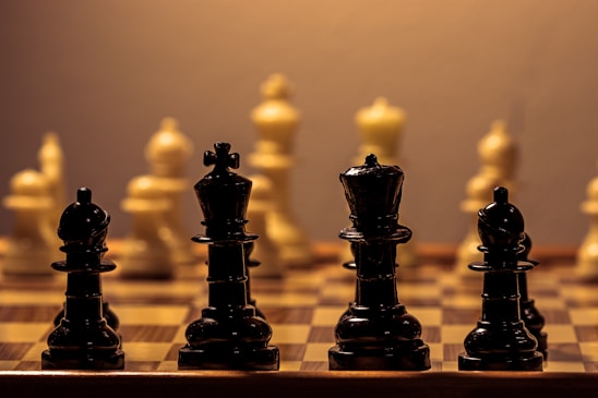 A close-up view of a chessboard highlighting the black chess pieces in the foreground with the white pieces in soft focus in the background. The black pieces, including the king and queen, are prominently displayed with a warm, ambient light casting soft shadows, creating a contemplative and strategic atmosphere.