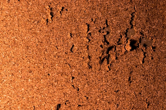 A textured surface of brown cork material, featuring an irregular, rough pattern with scattered dark indentations.