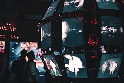 An array of screens in a dimly lit environment displays grainy, monochrome images of people. Each screen shows different scenes or angles, creating a fragmented and electronic aesthetic. The background contains colorful abstract graphics, contributing to a visually complex atmosphere. Silhouetted figures can be seen observing the display.