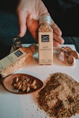 A hand holds a bottle of granulated garlic labeled 'Ajo Granulado' from 'Sho Premium'. Next to it, there is another bottle lying on its side and a wooden spoon containing garlic cloves. Some loose granulated garlic is spread on a white surface near garlic bulbs.