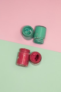 Two small jars of paint are positioned on a split background, one half pink and the other half green. The jars contain thick, colorful paint, with one jar containing red paint and the other containing teal paint. Both jars are open, showcasing the paint's texture.