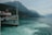 A scenic lake with a large white tour boat filled with passengers on the left foreground. The boat has decorative elements and a flagpole, with mountains covered in greenery in the background under a cloudy sky. The water near the boat is a vibrant turquoise, creating a contrast with the forested landscape.