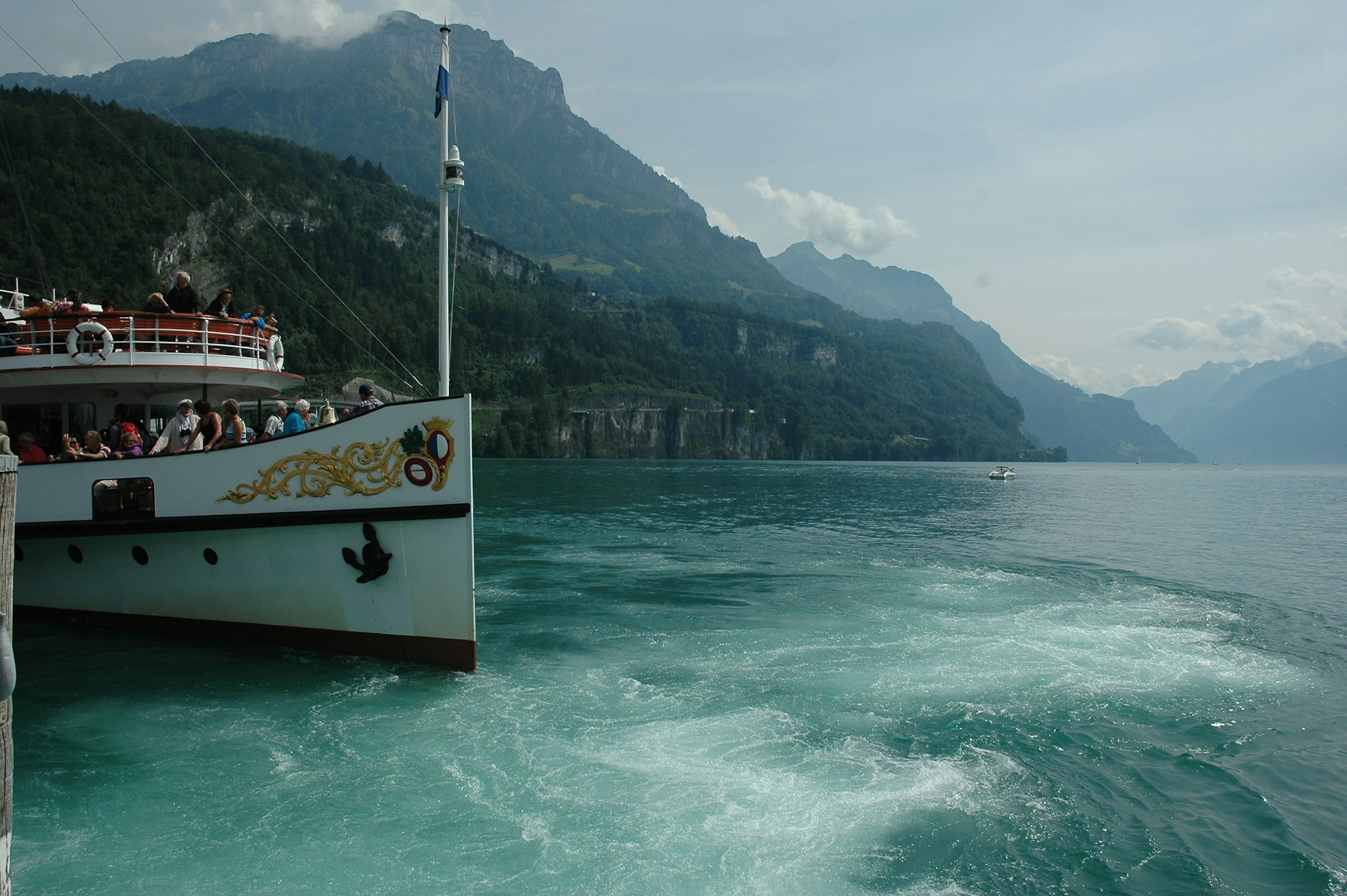 A scenic lake with a large white tour boat filled with passengers on the left foreground. The boat has decorative elements and a flagpole, with mountains covered in greenery in the background under a cloudy sky. The water near the boat is a vibrant turquoise, creating a contrast with the forested landscape.
