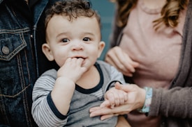 A young child with curly hair wearing a striped shirt holds fingers to their mouth while being gently held by an adult. The adult's hands are visible, one supporting the child and the other holding the child's hand. The background includes portions of adult clothing, featuring denim and a soft sweater.