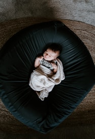 A newborn baby is peacefully lying on a dark, rounded cushion with a textured blanket partially covering them. The baby is wearing a onesie that says 'Hello World' and is surrounded by a warm, serene environment.