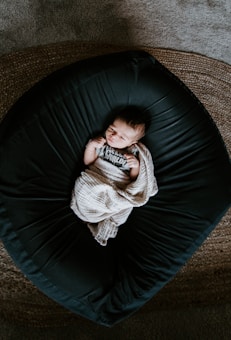 A newborn baby is peacefully lying on a dark, rounded cushion with a textured blanket partially covering them. The baby is wearing a onesie that says 'Hello World' and is surrounded by a warm, serene environment.