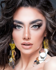 A close-up portrait of a person with striking blue eyes, full, arched eyebrows, and long, voluminous eyelashes. The makeup is sophisticated, highlighting the eyes and lips, with a soft nude lipstick. The hair is styled in loose, wavy curls. Large, shiny, circular earrings with gold detailing add a glamorous touch.